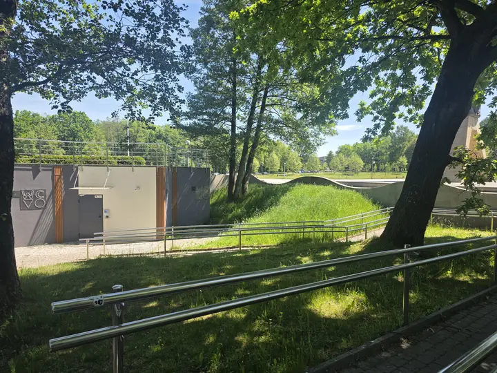 Widok alei parkowej, po lewej toalety miejskie, po prawej fragment pomnika
