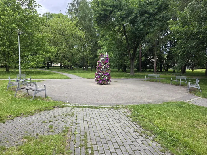Kolejny placyk w parku z ławkami a pośrodku niego w sezonie wiosenno-letnim kwietnik