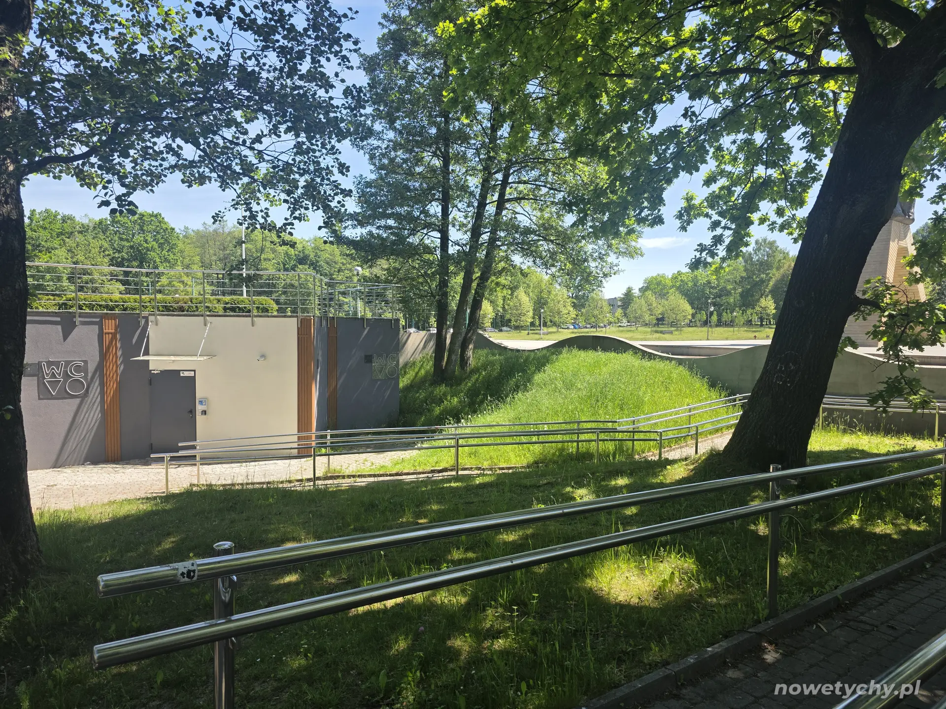 Widok alei parkowej, po lewej toalety miejskie, po prawej fragment pomnika