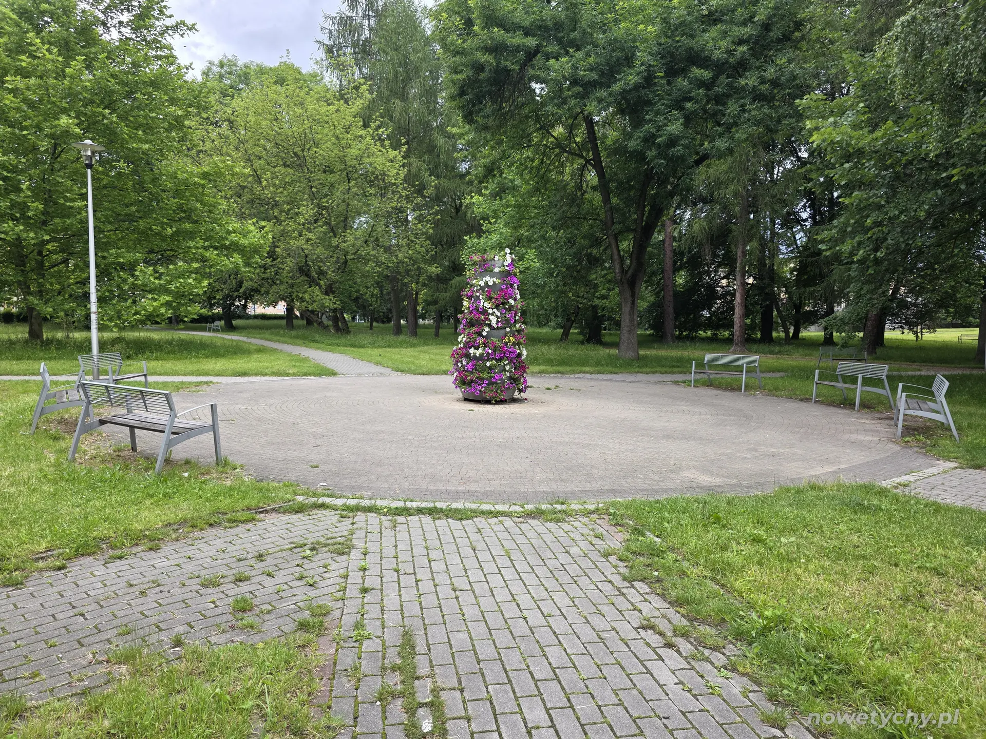 Kolejny placyk w parku z ławkami a pośrodku niego w sezonie wiosenno-letnim kwietnik